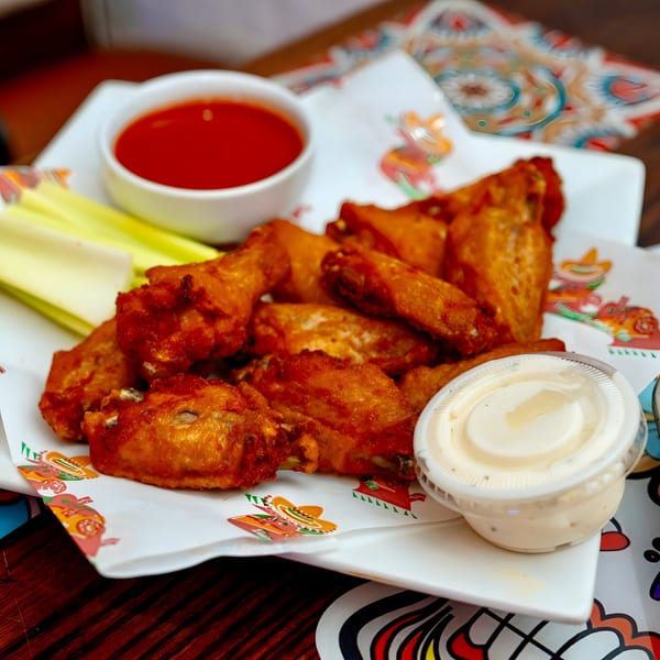 Crispy chicken wings with celery sticks, red hot sauce, and ranch dressing on a Pedros Tacos plate.