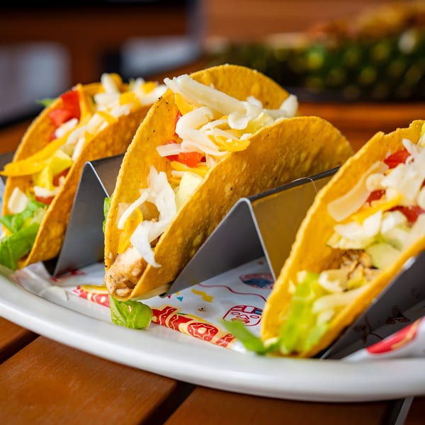 Three Pedros Tacos with chicken, lettuce, cheese, and tomatoes in a metal holder on a white plate.