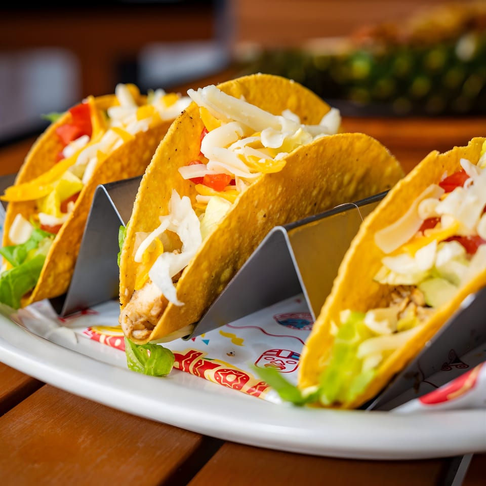 Three Pedros Tacos with chicken, lettuce, cheese, and tomatoes in a metal holder on a white plate.
