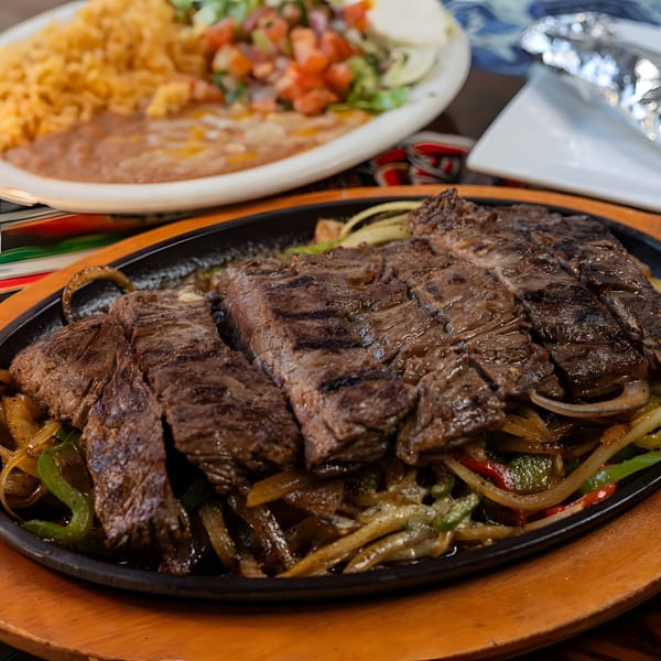 Sizzling steak fajitas with grilled onions and peppers, with rice and beans in the background.