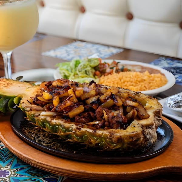 Grilled pineapple half filled with steak, onions, and pineapple chunks, served with rice and salad.