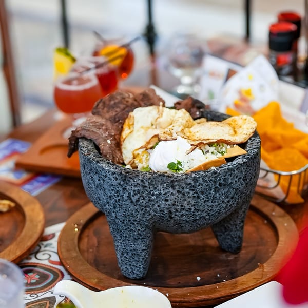 Molcajete dish with steak, tortillas, and sour cream, alongside drinks and chips at Pedros Tacos & Tequila Bar.